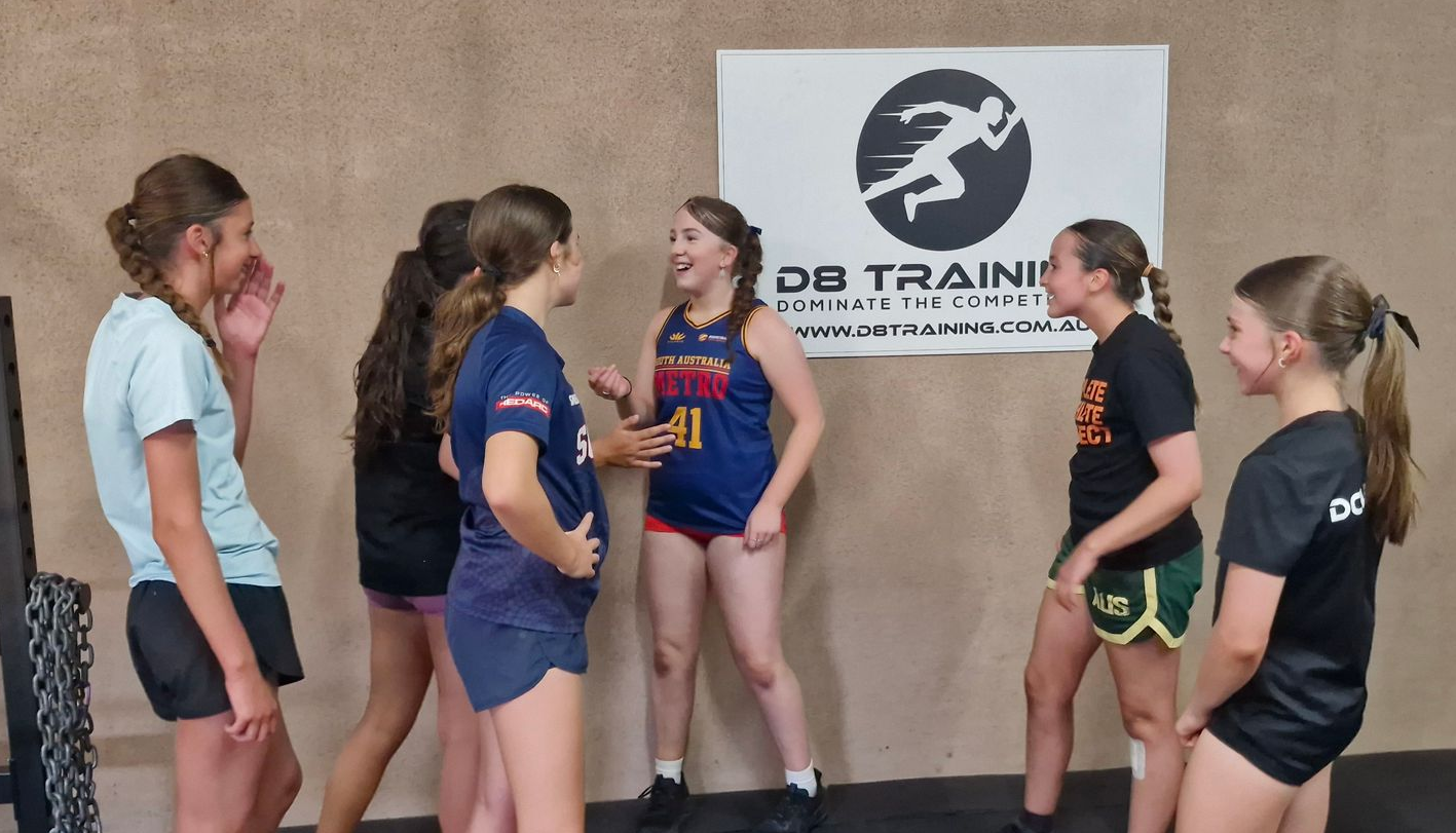 Five athletes in training gear stand talking in front of a wall sign at a gym.