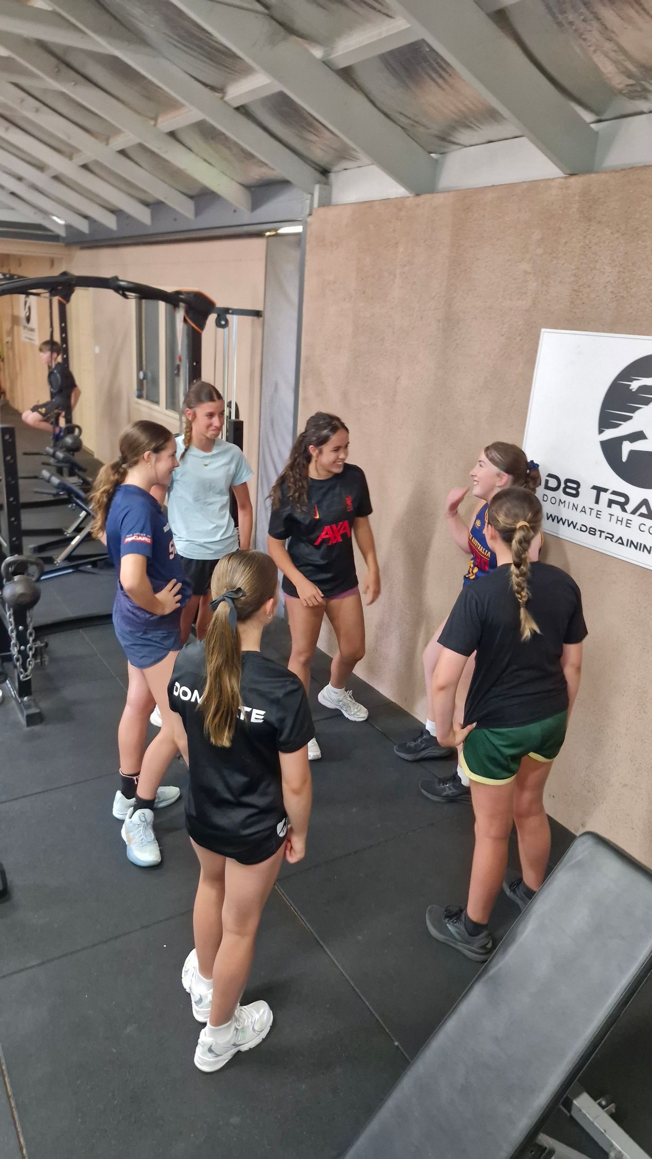 Group of people in workout clothes talking in a gym near a wall with a logo.