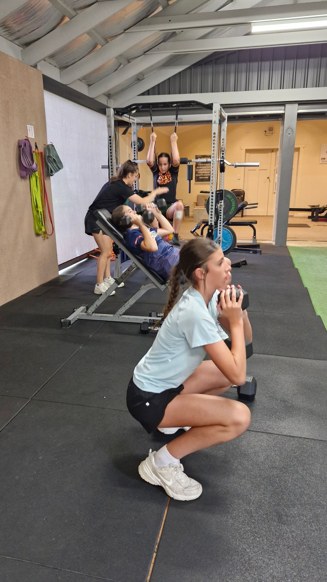 People exercising in a gym, one person squatting in the foreground near weight racks.