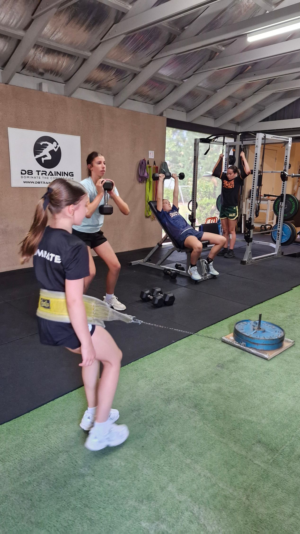 Group fitness class in an open-air gym, with people exercising on mats and equipment under a wooden roof.