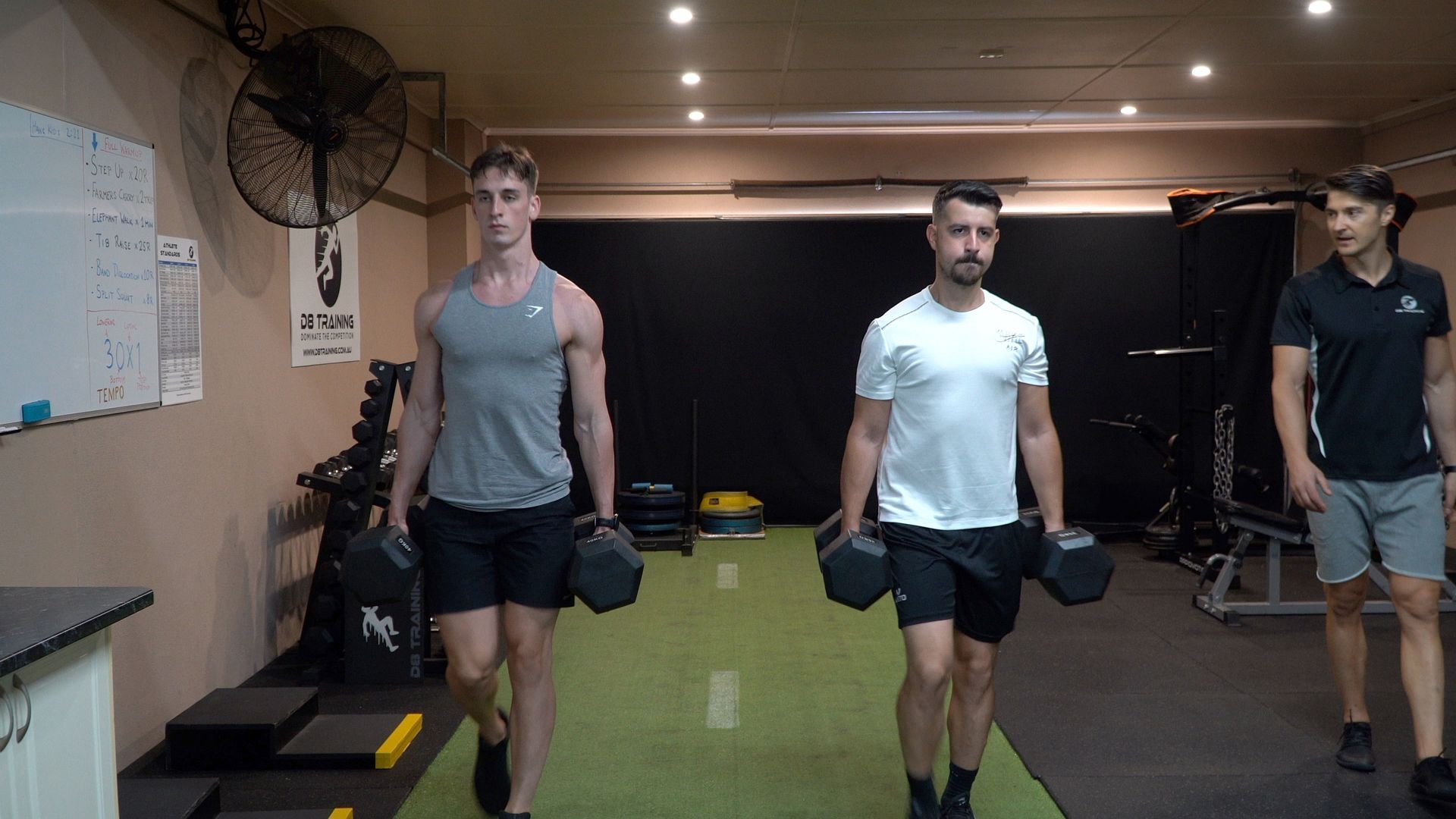 Three men walking with dumbbells in a gym. One in a gray tank top, one in white tee, and one in black polo. Green and black floor.