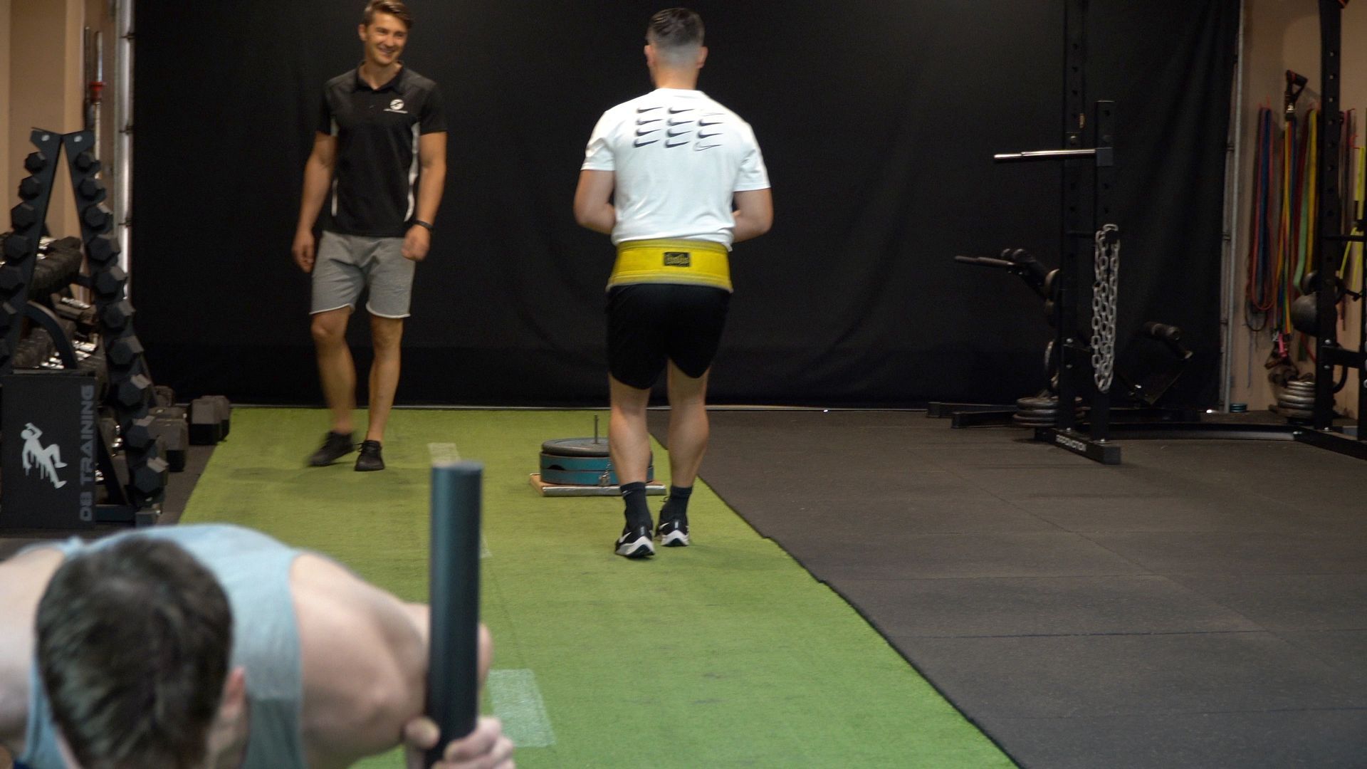 Man walking with a weighted belt in a gym. Another man observes. A third person is performing an exercise.