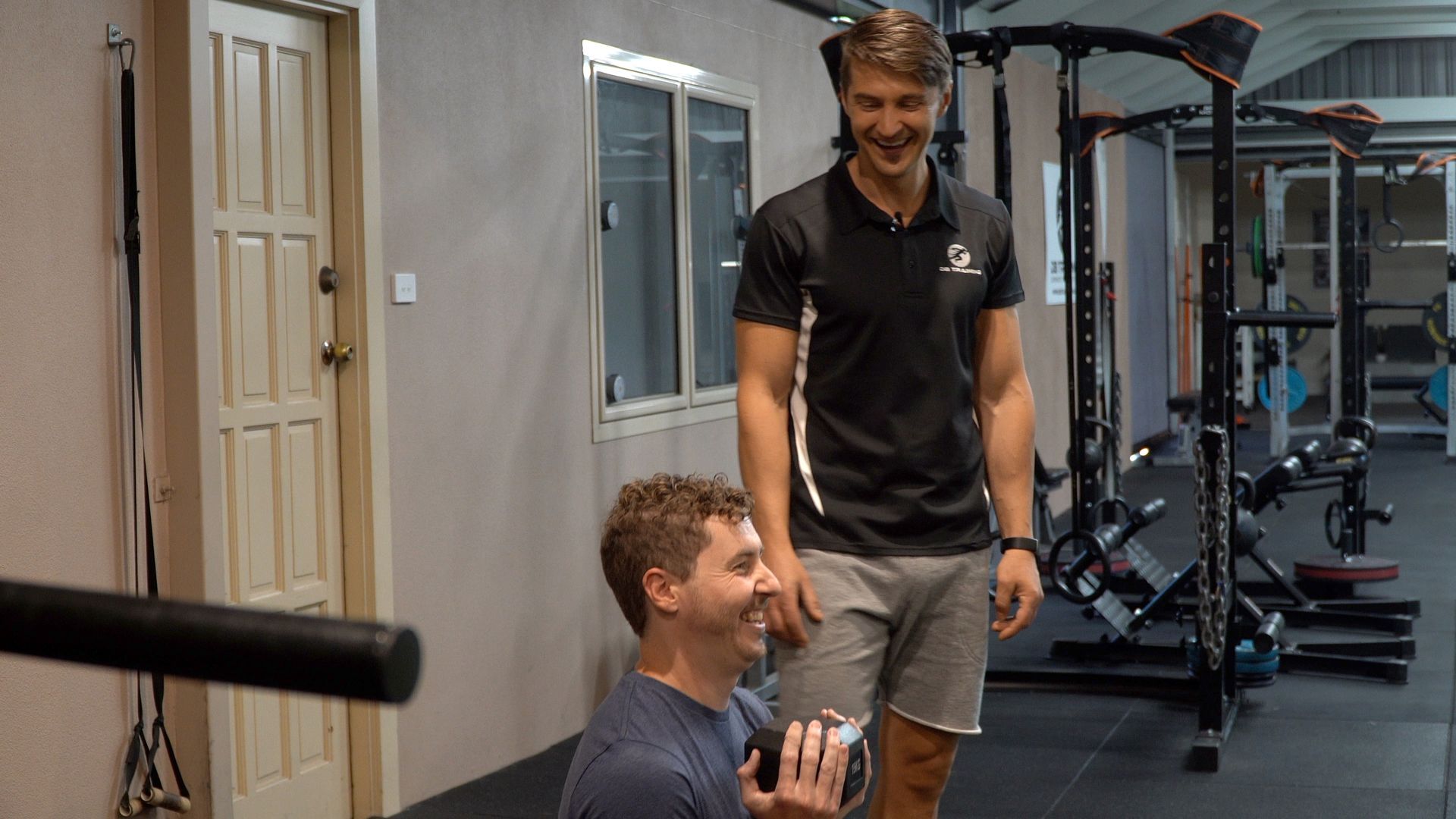 Two people chatting in a gym, one standing and smiling while the other sits holding a drink.