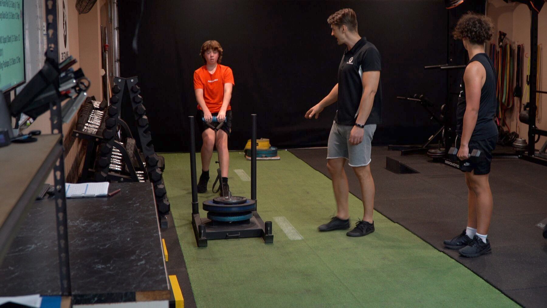 People in a gym watching one person push a sled on a green turf lane