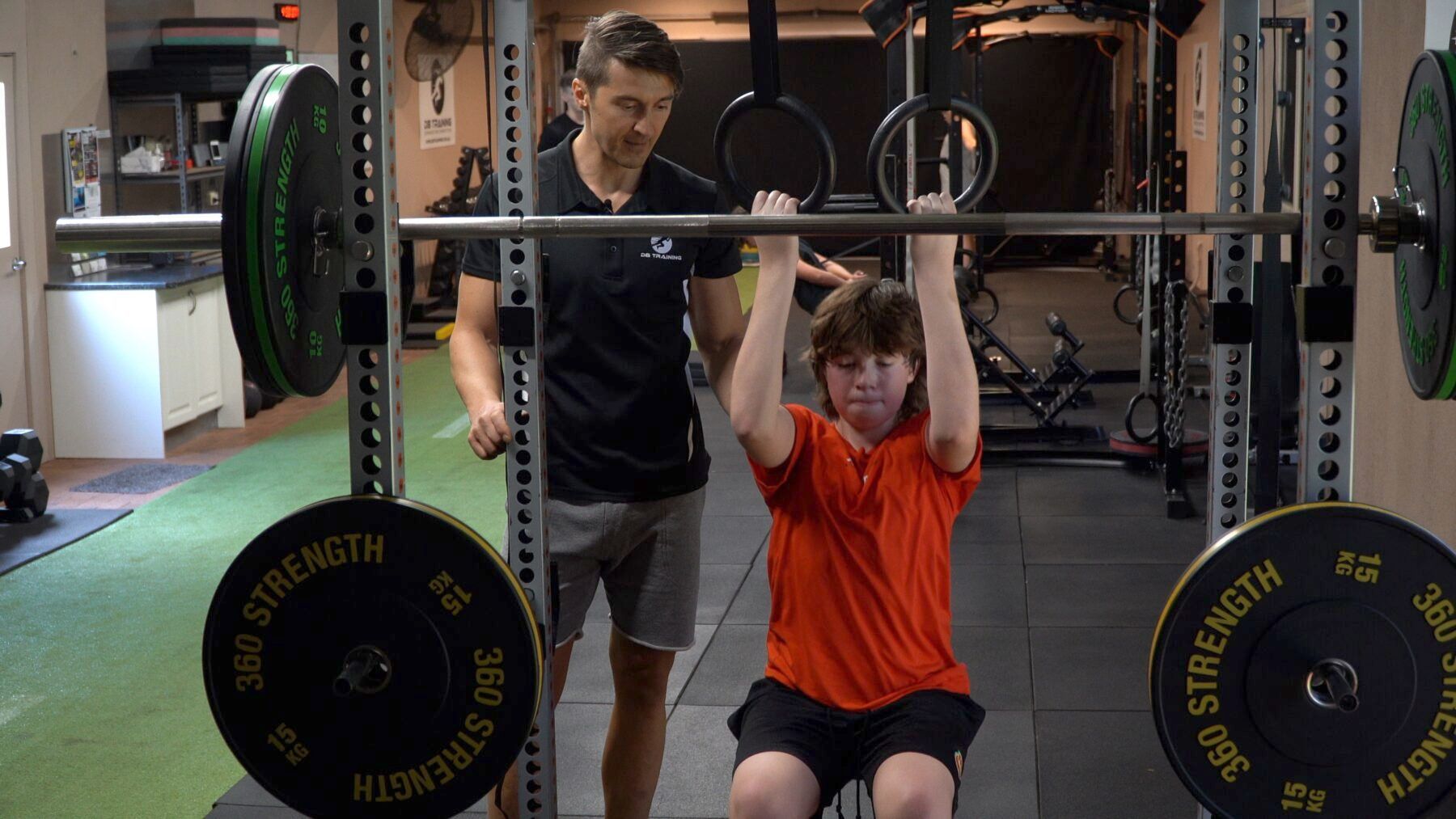 Man assisting a seated person in an orange shirt lifting a barbell in a gym