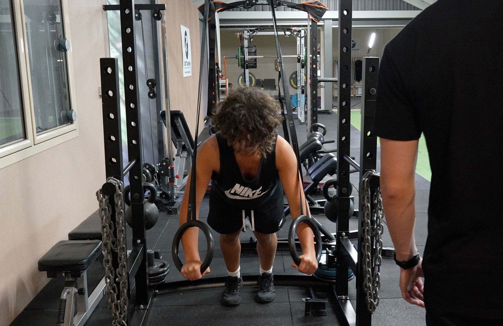 Person lifting a barbell in a gym, with a trainer standing nearby.