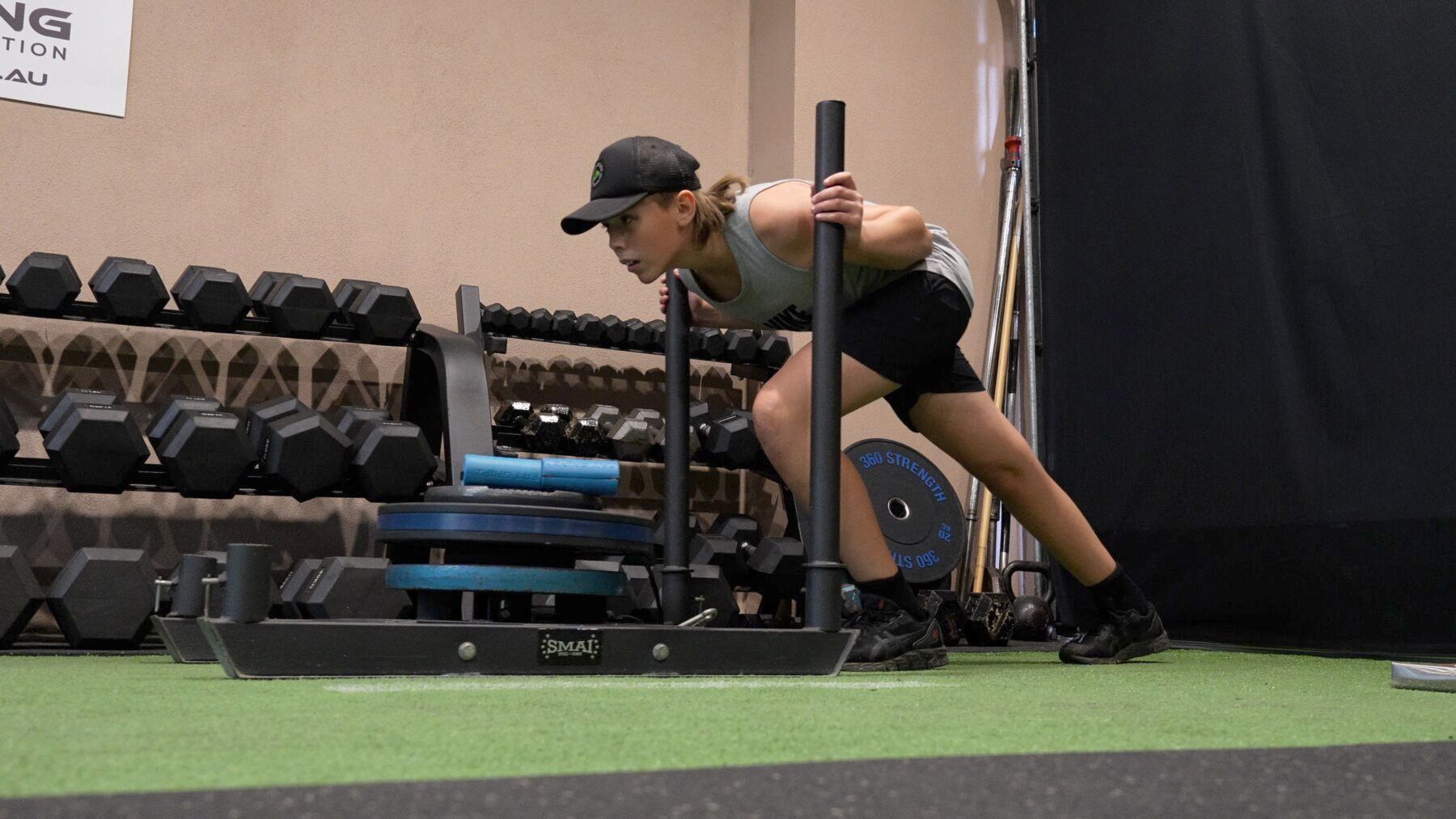Person pushing a weighted sled on indoor turf near dumbbells.