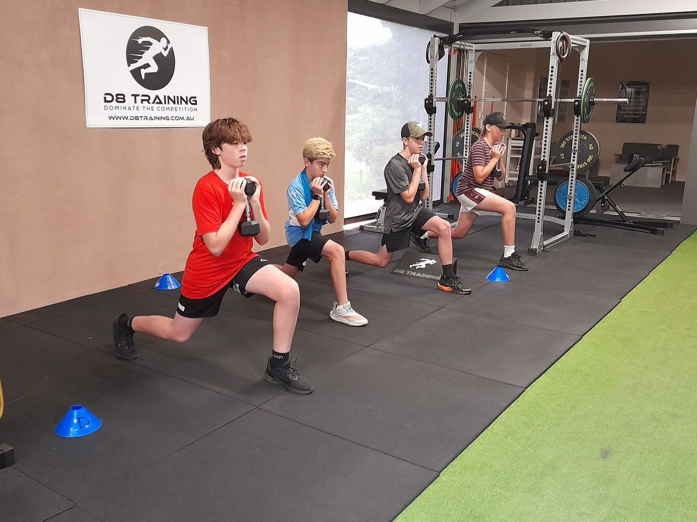 People exercising in a gym, lunging with dumbbells on black mats near blue cones