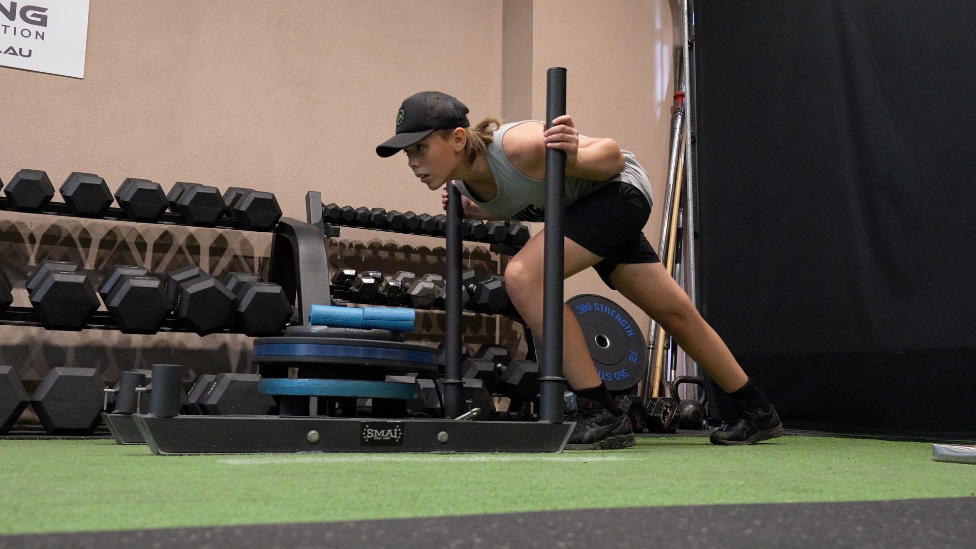 Person pushing a weighted sled on indoor turf beside dumbbells in a gym