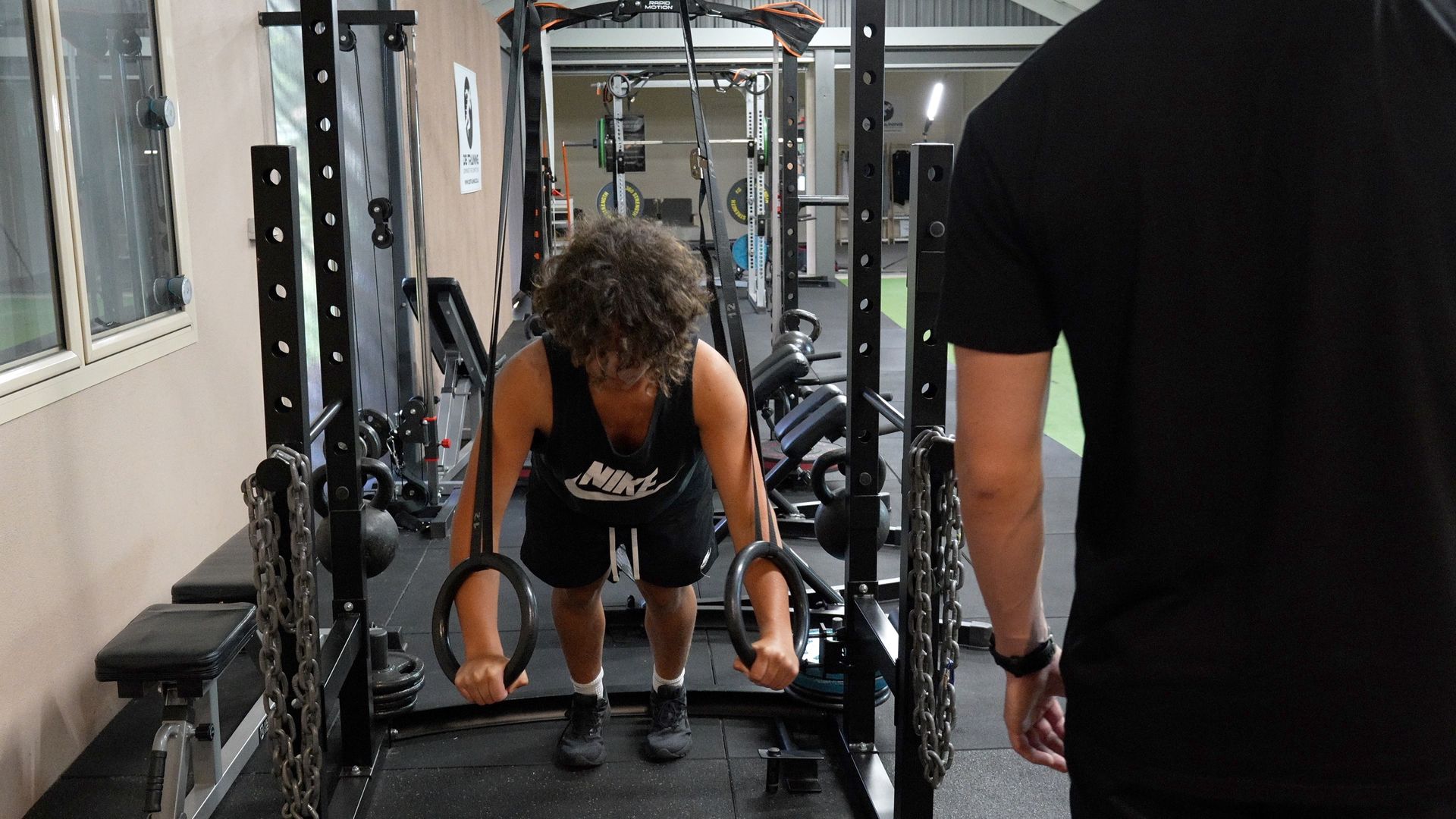 Person doing a bent-over dumbbell exercise in a gym, with another person nearby.