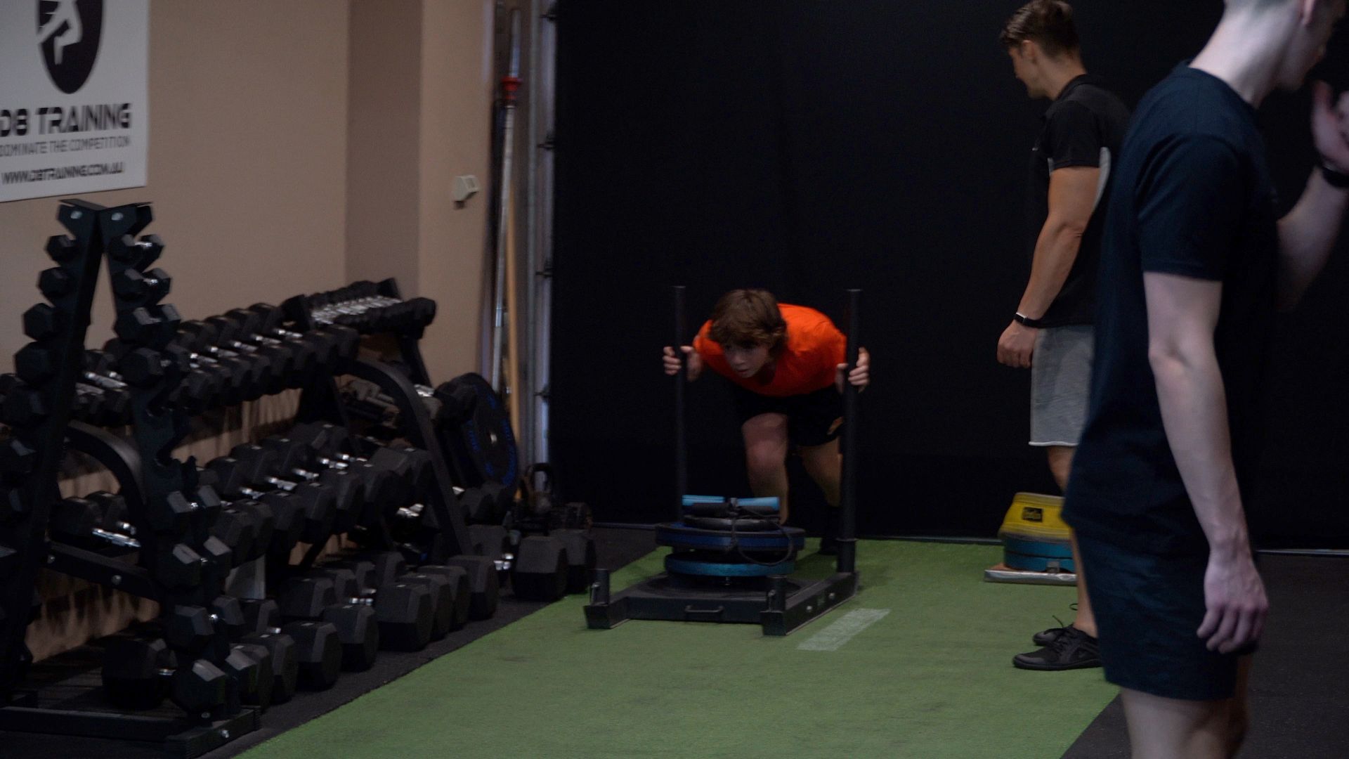 People training in a gym beside dumbbells, with one person bent over a sled on turf.