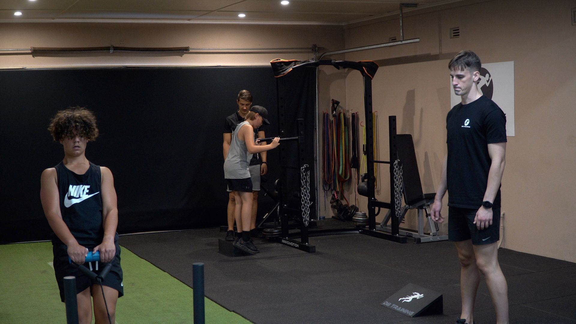 People exercising in a gym, with one holding dumbbells and others using equipment in the background.