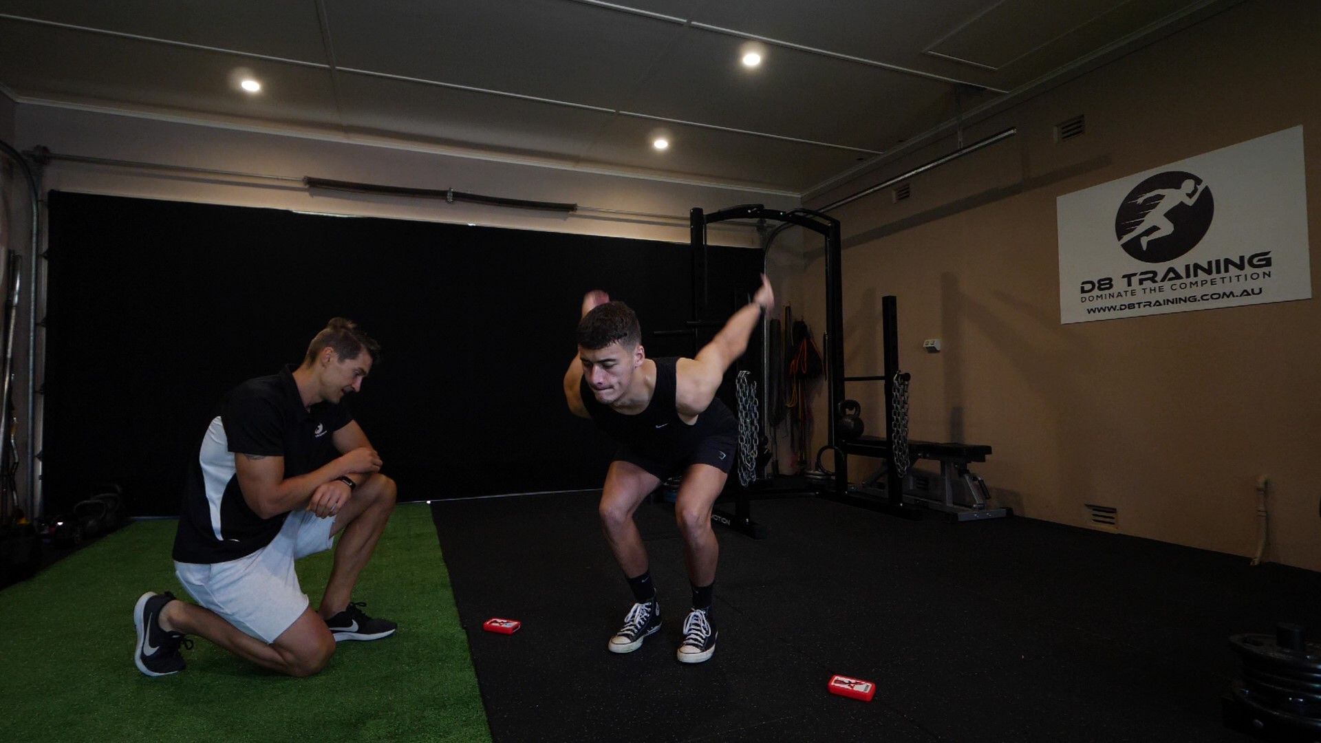 Man performing squat exercise, watched by coach. Gym setting with black backdrop and sports equipment.