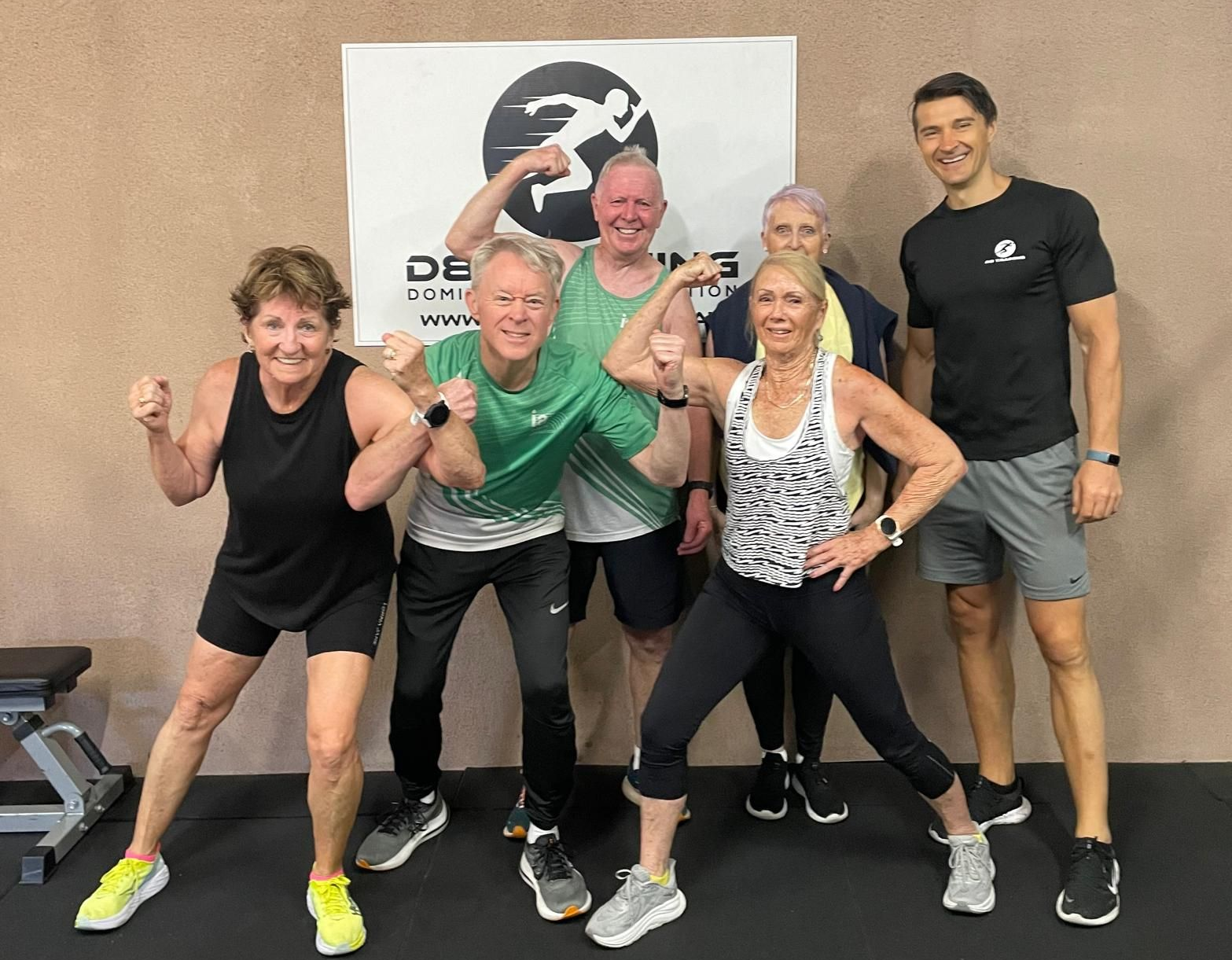 Five people posing and flexing in a gym, with workout equipment and a fitness logo banner behind them