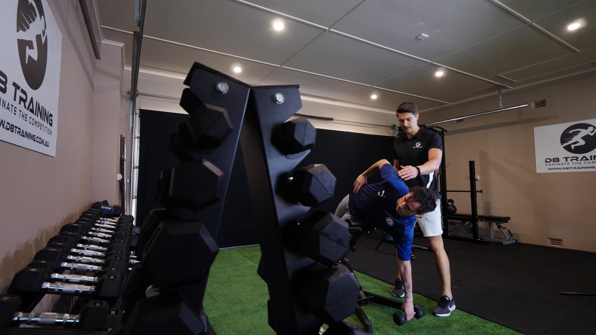 Man assisting another man in a gym; the man bends over to lift a dumbbell.