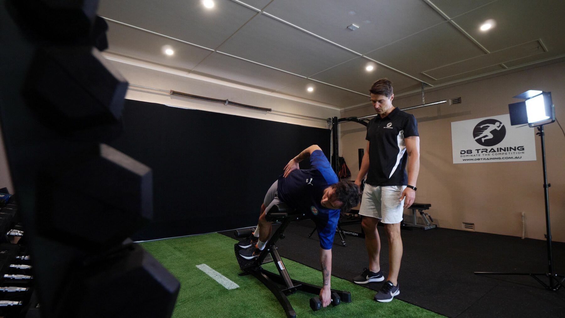 Two people on indoor golf turf, one bending to adjust equipment while the other stands nearby.