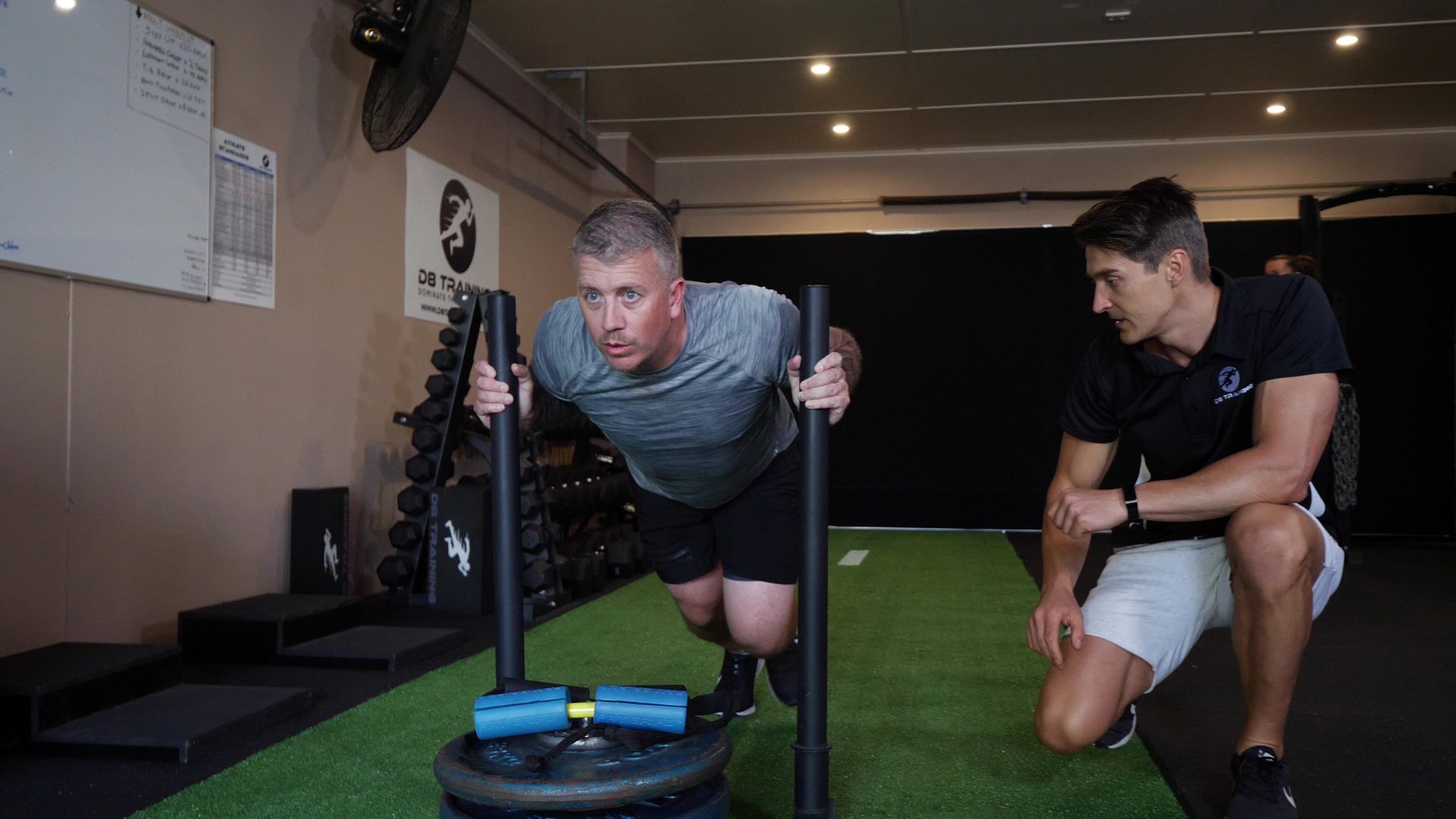 Two men training with a weighted sled in a gym, one pushing hard while the other kneels beside him.