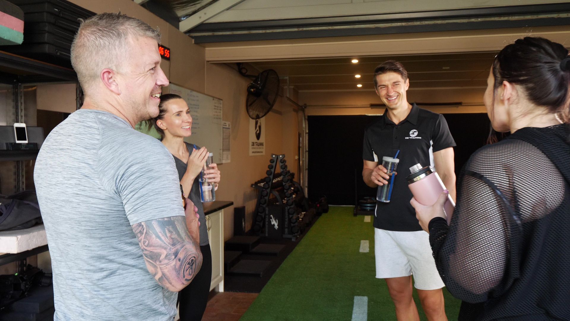 People chatting in a gym hallway beside turf, one holding a drink, with exercise equipment in the background