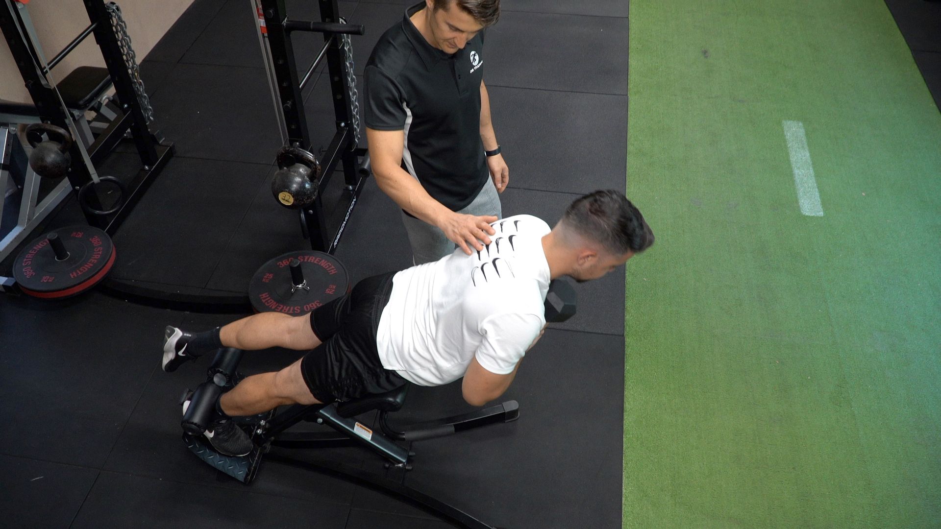 Man doing back extension exercise, instructor assisting. Black gym setting, green mat.