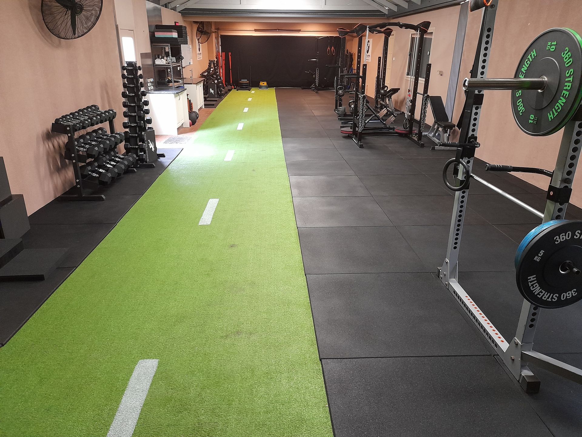 Empty gym with green turf lane, weight racks, and exercise mats on the floor.