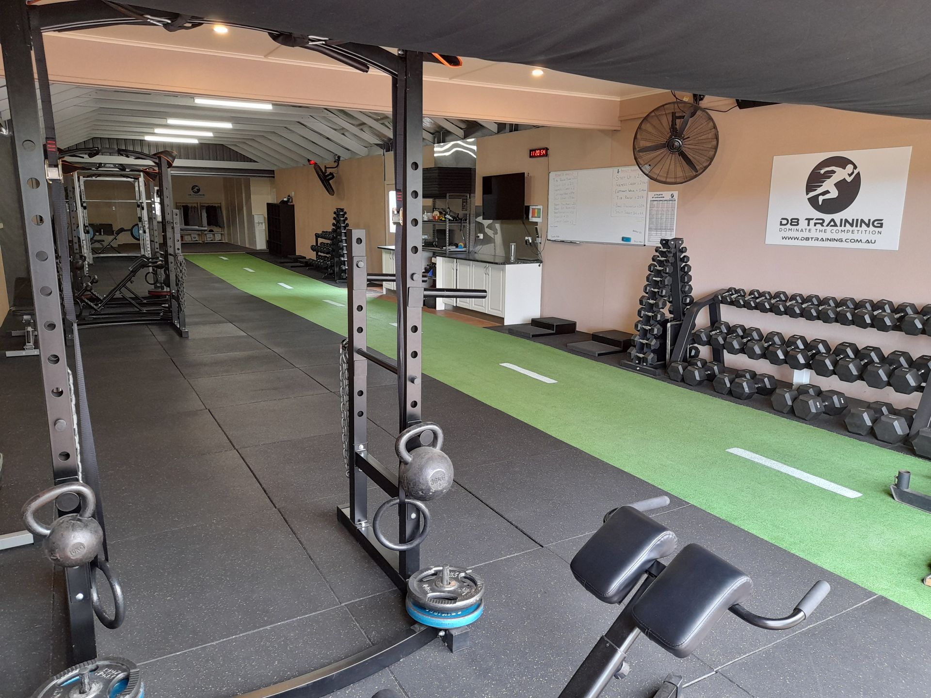 Gym interior with kettlebells, dumbbells, bench, mirrored wall, and turf lane under ceiling lights