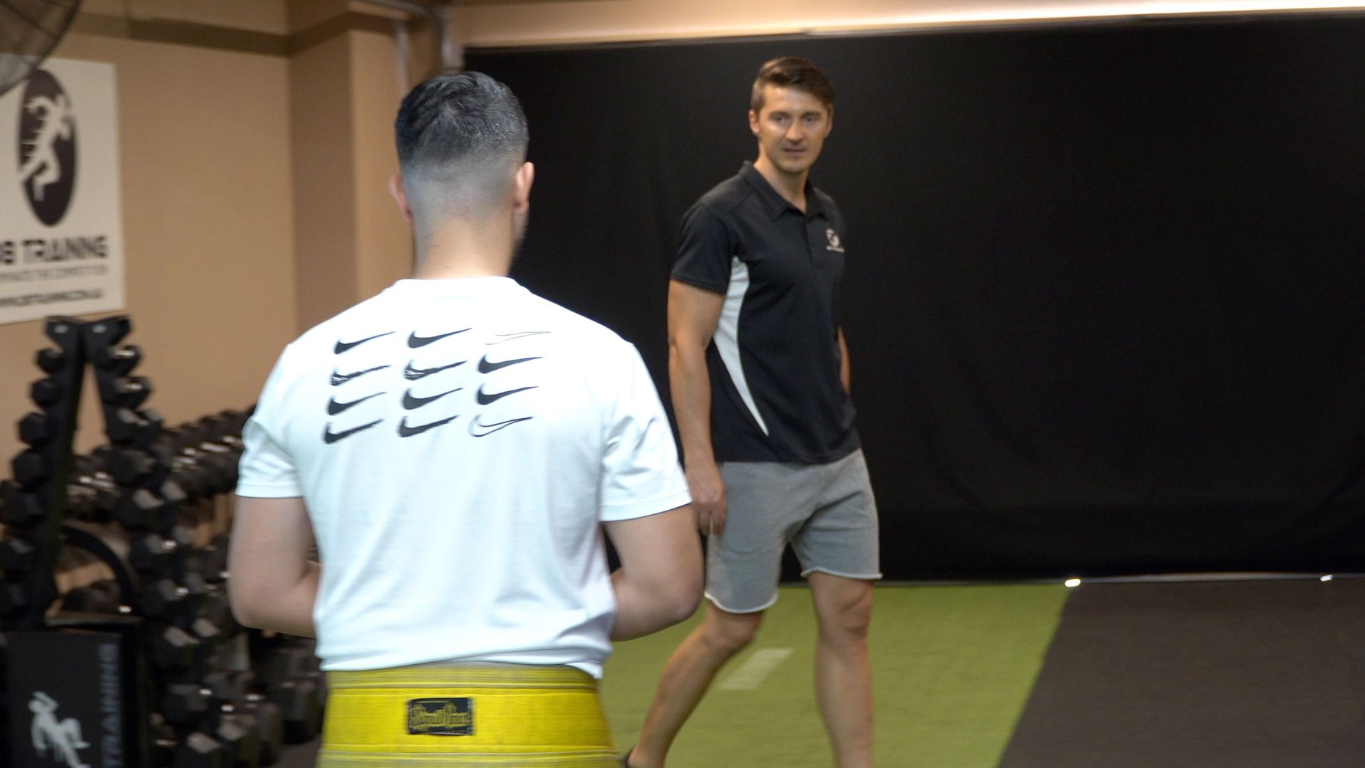 Two men training in a gym, one in a white shirt and yellow shorts facing a coach in black.