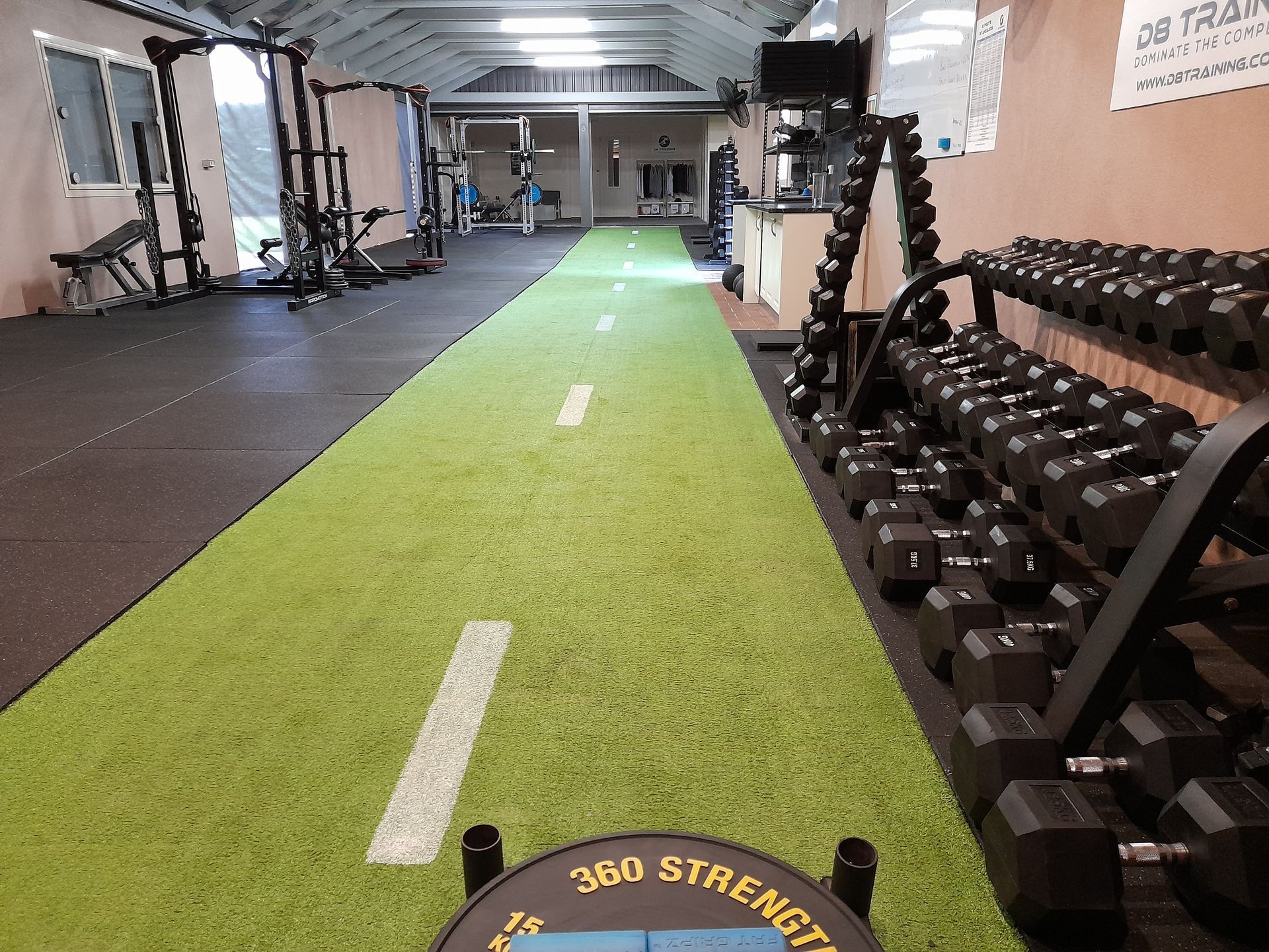Indoor gym lane with green turf and rows of dumbbells on the right