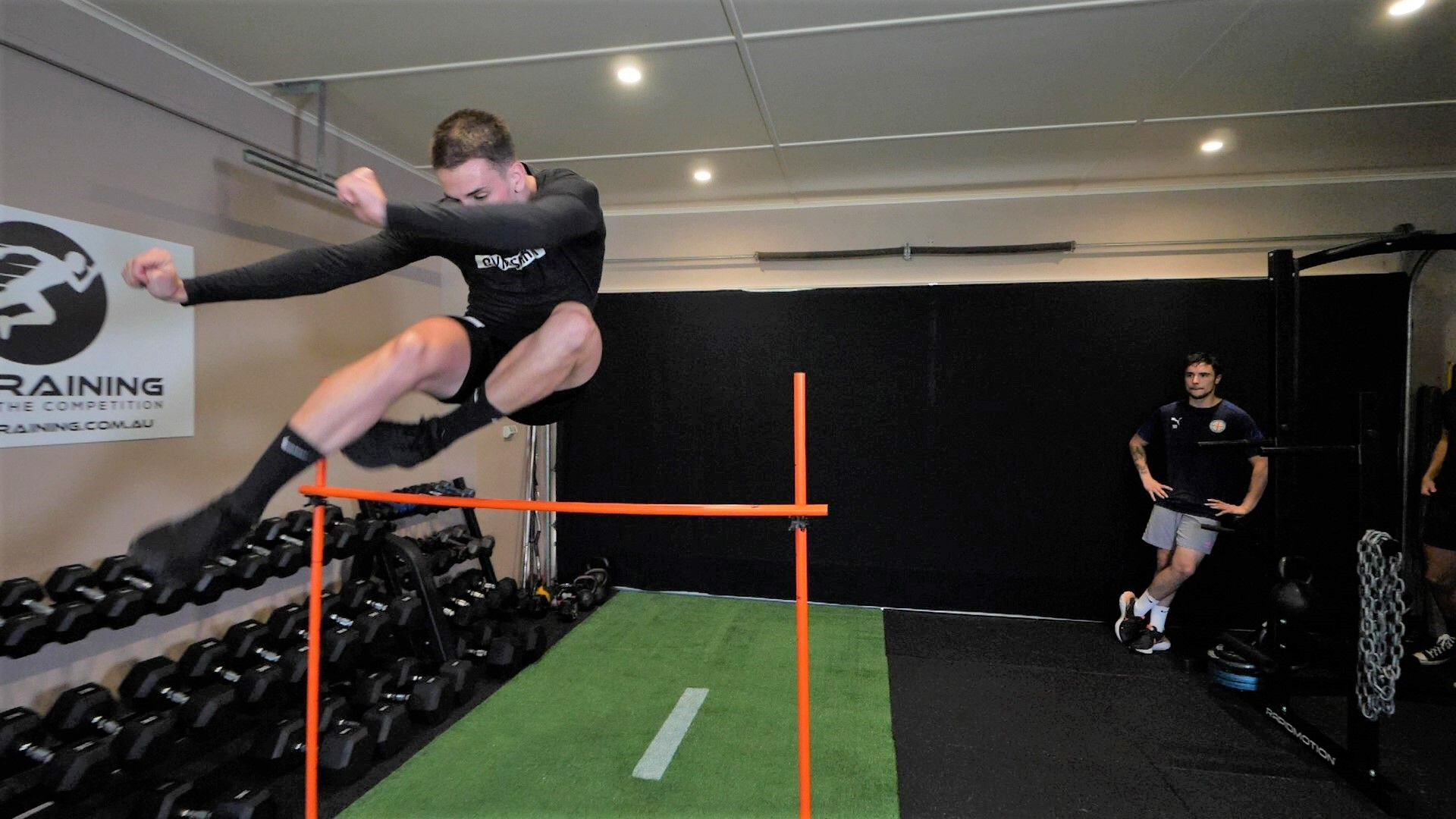 Man jumping over a hurdle in a gym, arm extended, black clothing, green mat.