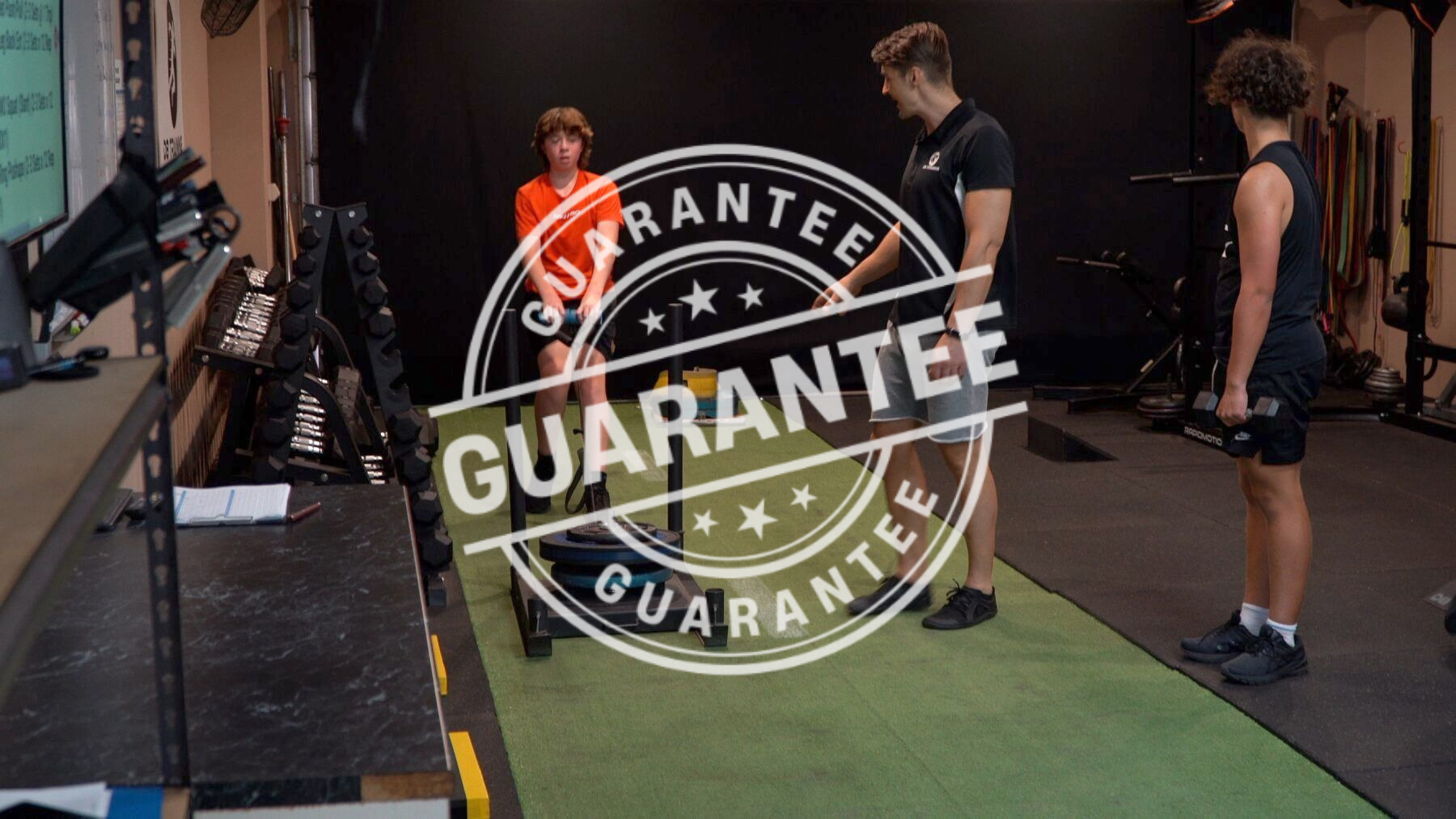 Three people standing in a gym beside a barbell, with a large “GUARANTEE” stamp overlay
