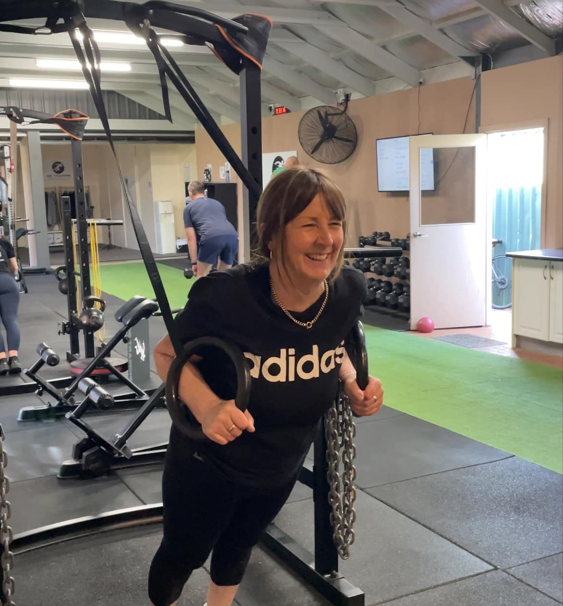 Woman smiling on gym cable machine, wearing black Adidas shirt in a workout space.