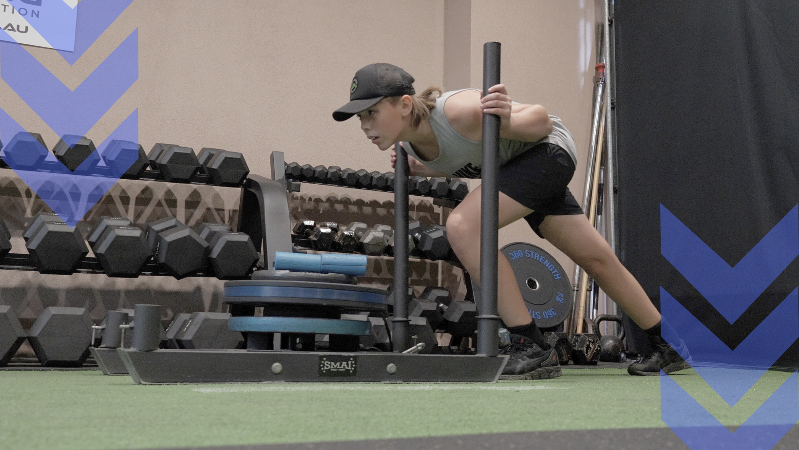 Woman in gym pushes weighted sled; black and blue weights; holding handles; green mat; wearing hat and shorts.