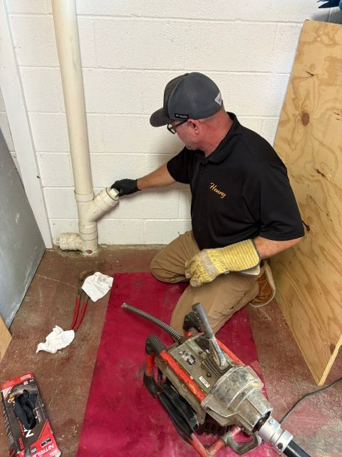 Technician kneeling by a wall pipe, using tools on a basement floor with a red mat.