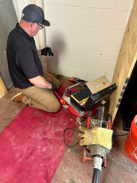 Technician kneeling beside an open wall, using tools to inspect wiring near a red carpet and equipment case