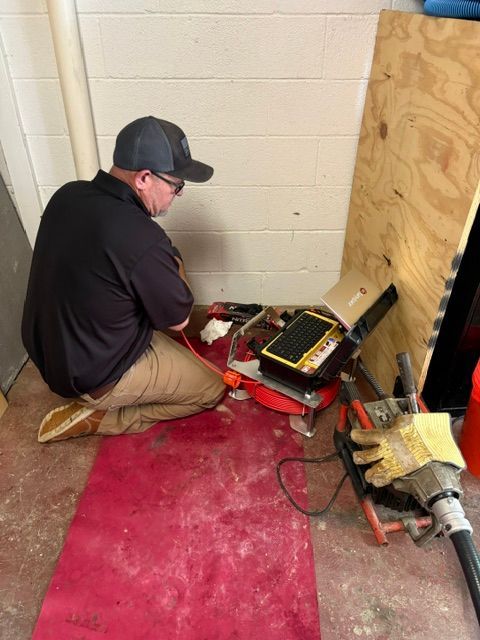 Worker kneeling by a red machine in a utility room, examining tools and wires on the floor.