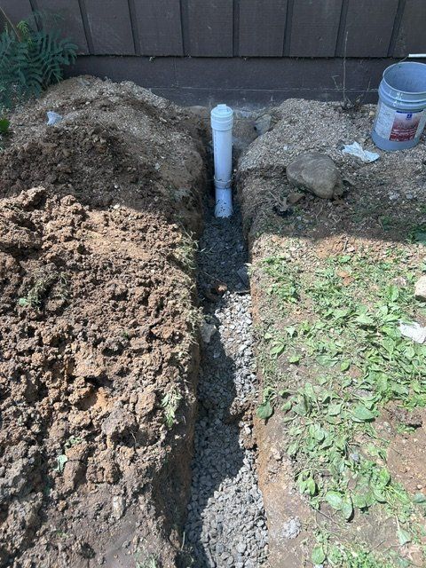 Open trench in a yard with exposed pipe and dirt piles near a house foundation