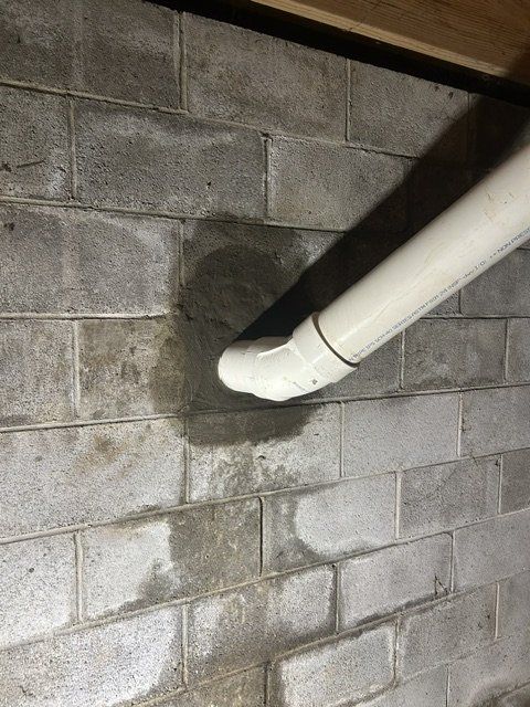 White pipe elbow running into a gray cinder block wall, with a damp stain beneath it.