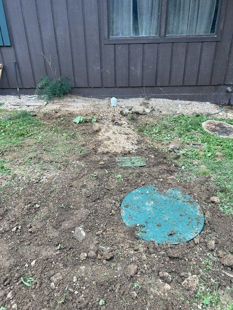 Dirt patch beside a dark wooden building, with grass and a blue circular cover in the foreground.