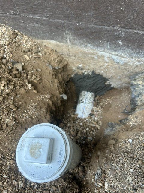 Utility excavation with a white pipe cap and exposed pipe in dirt beside a concrete wall