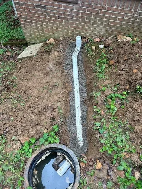 White pipe running through a muddy yard to a brick wall, with a round utility box in the foreground