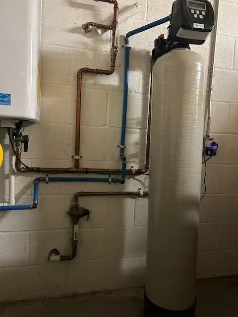 Water softener and plumbing pipes in a utility room against a cinderblock wall.