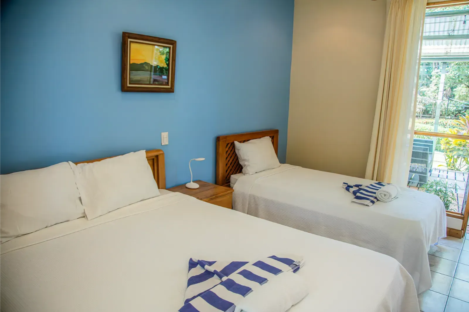 A bedroom with two beds, blue wall, painting, and balcony view.