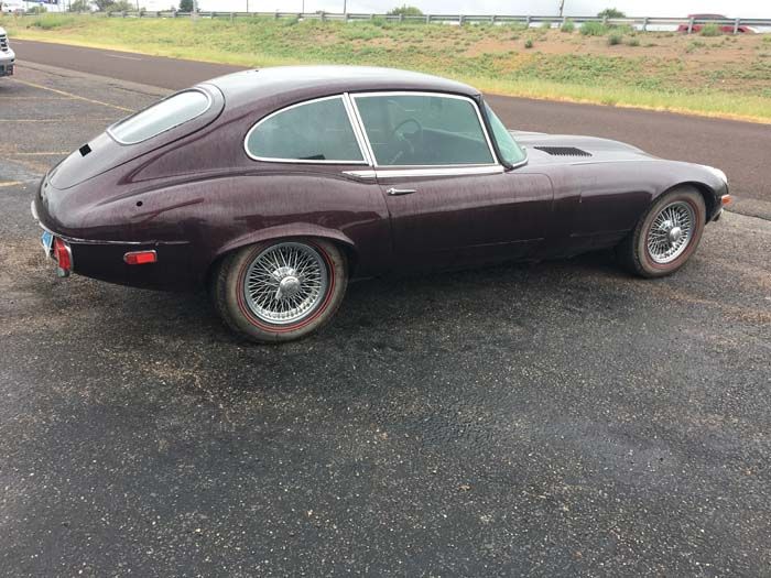 Burgundy Jaguar E-Type coupe parked on asphalt, silver wire wheels, tinted windows, setting is outdoors.