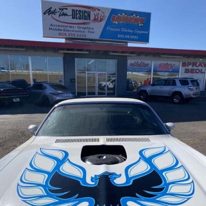 White classic car with blue bird graphic on the hood parked in front of a shop.