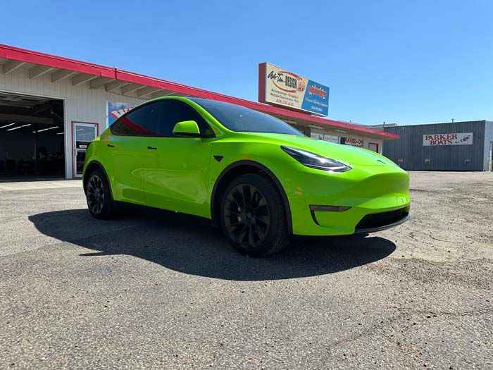 Bright green Tesla Model Y parked in front of a shop on a sunny day. Black wheels, tinted windows.