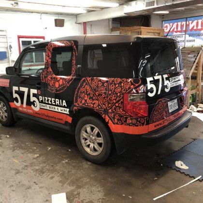 Black vehicle with pizza-themed wrap advertising 575 Pizzeria. Orange accents. Parked in garage.