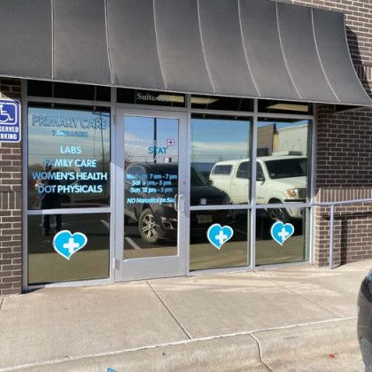 Exterior of a primary care clinic with glass windows, displaying services, hours, and heart logos.