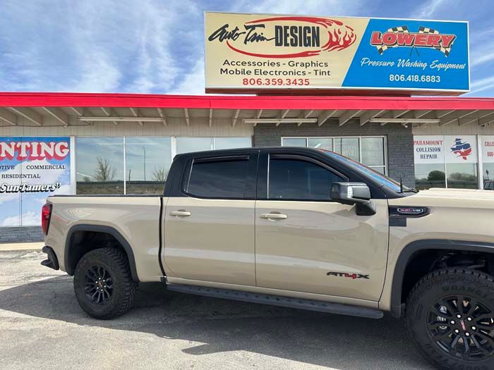 Tan GMC truck parked outside a shop with signage for auto tint, graphics, and accessories.