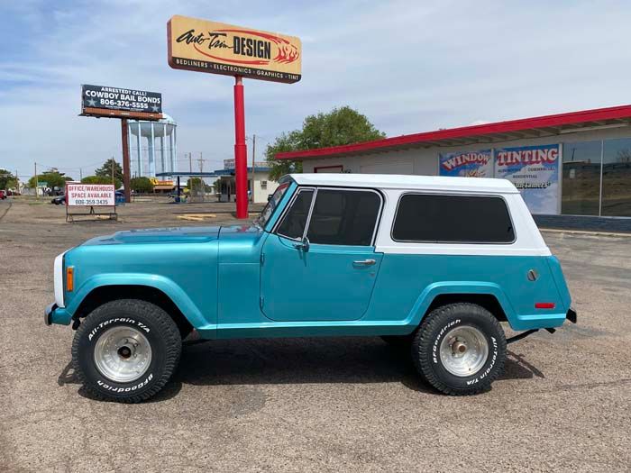 Teal and white vintage Jeep parked in front of a business with tinted windows and a red awning.