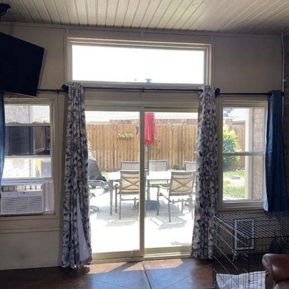 Sliding glass door to patio with table, chairs, and fence. Windows on either side, curtains, TV, and AC.