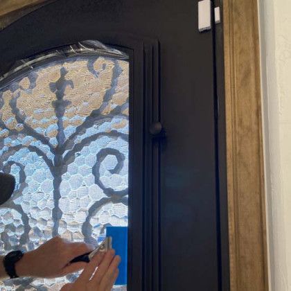 Person using a tool to apply something to a black door with decorative glass, indoors.